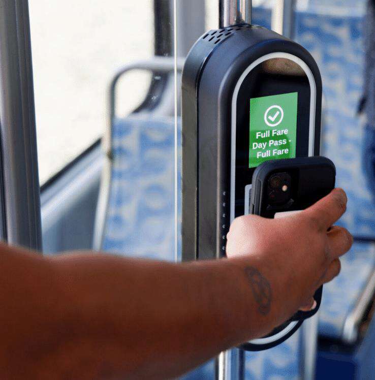 Passenger holding their phone up to a scanner to pay their bus fare. A green checkmark appears on the scanner, confirming payment.